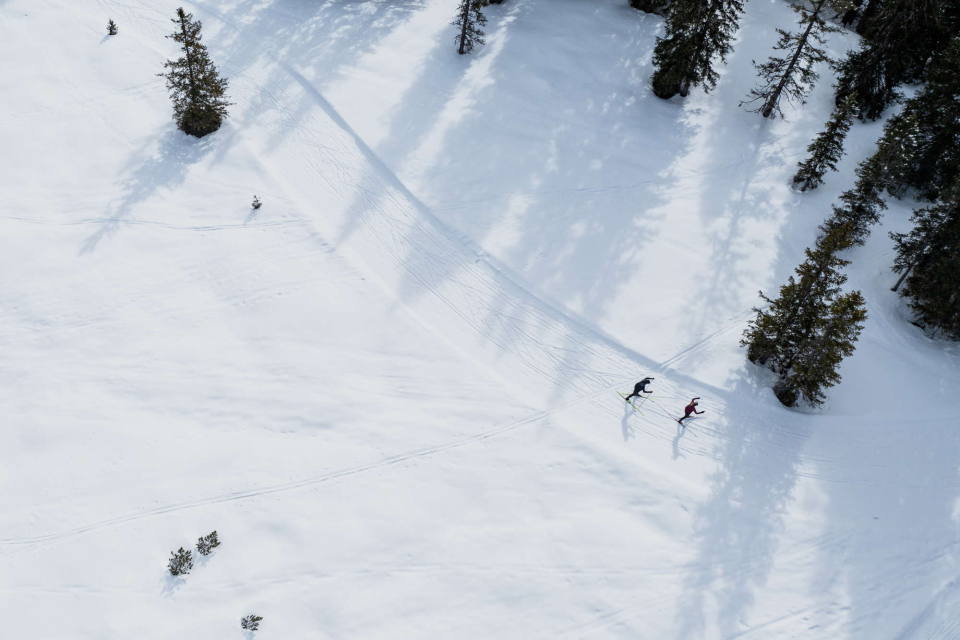Zwei Langläufer gleiten nebeneinander durch eine verschneite Waldlandschaft, aufgenommen aus der Vogelperspektive.
