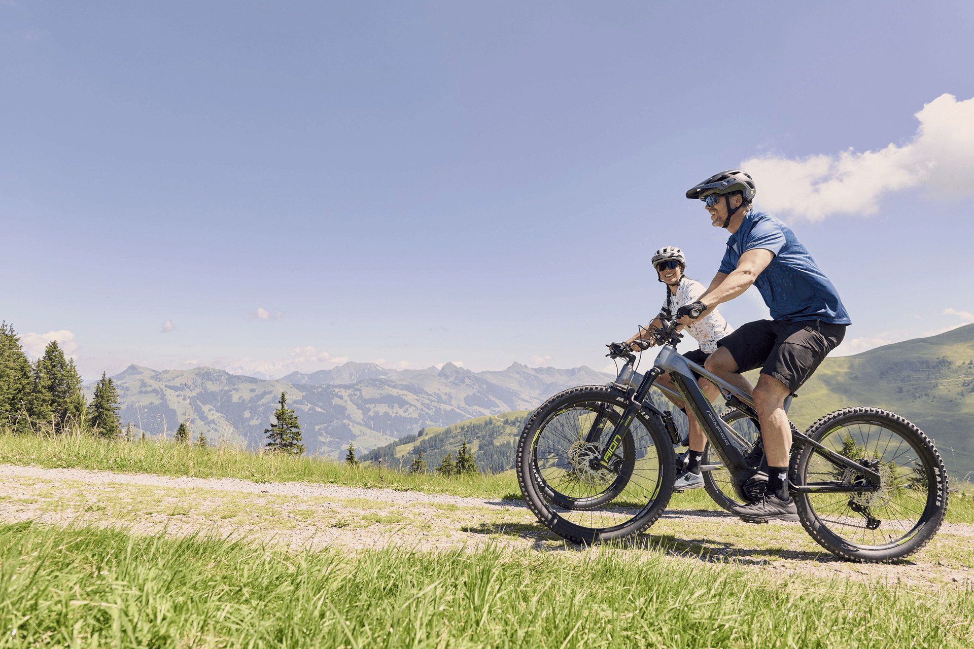 Zwei Radfahrer fahren mit Mountainbikes auf einem grasbewachsenen Weg mit malerischem Blick auf ferne Berge unter einem klaren blauen Himmel und vermitteln dabei Abenteuerlust und Freude.