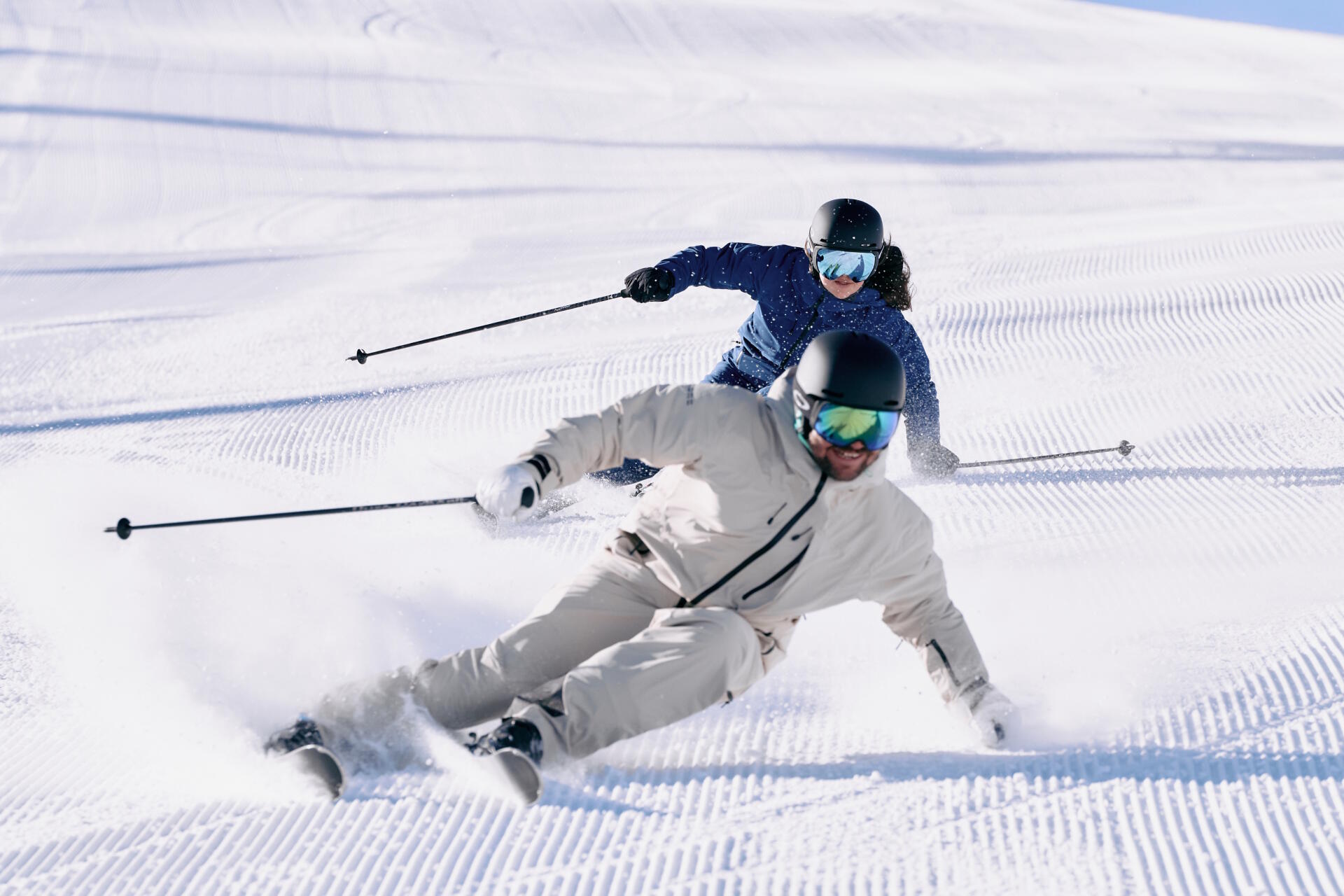 Zwei Skifahrer in hellem und blauem Outfit carven dynamisch parallel über eine frisch präparierte Skipiste.