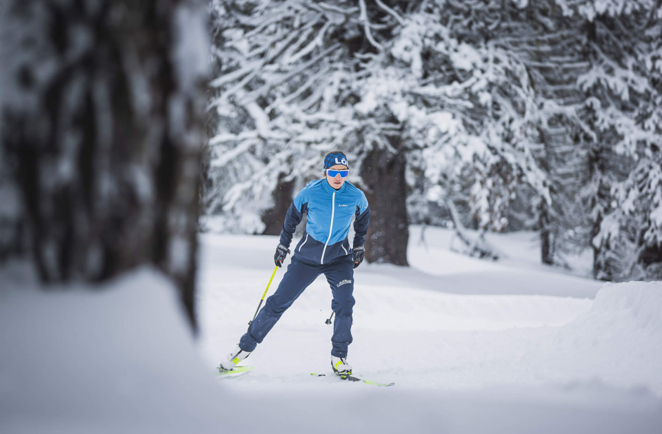 Cross-country skier in blue winter clothing skiing through a snow-covered forest with deep powder snow.