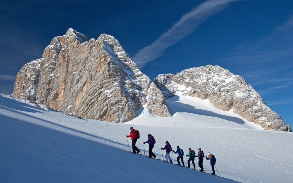 Zwei Skifahrer in grauen Outfits und Helmen stehen auf einem verschneiten Hang, halten ihre Skier in den Händen, während im Hintergrund ein strahlend blauer Himmel und ein Berggipfel zu sehen sind.