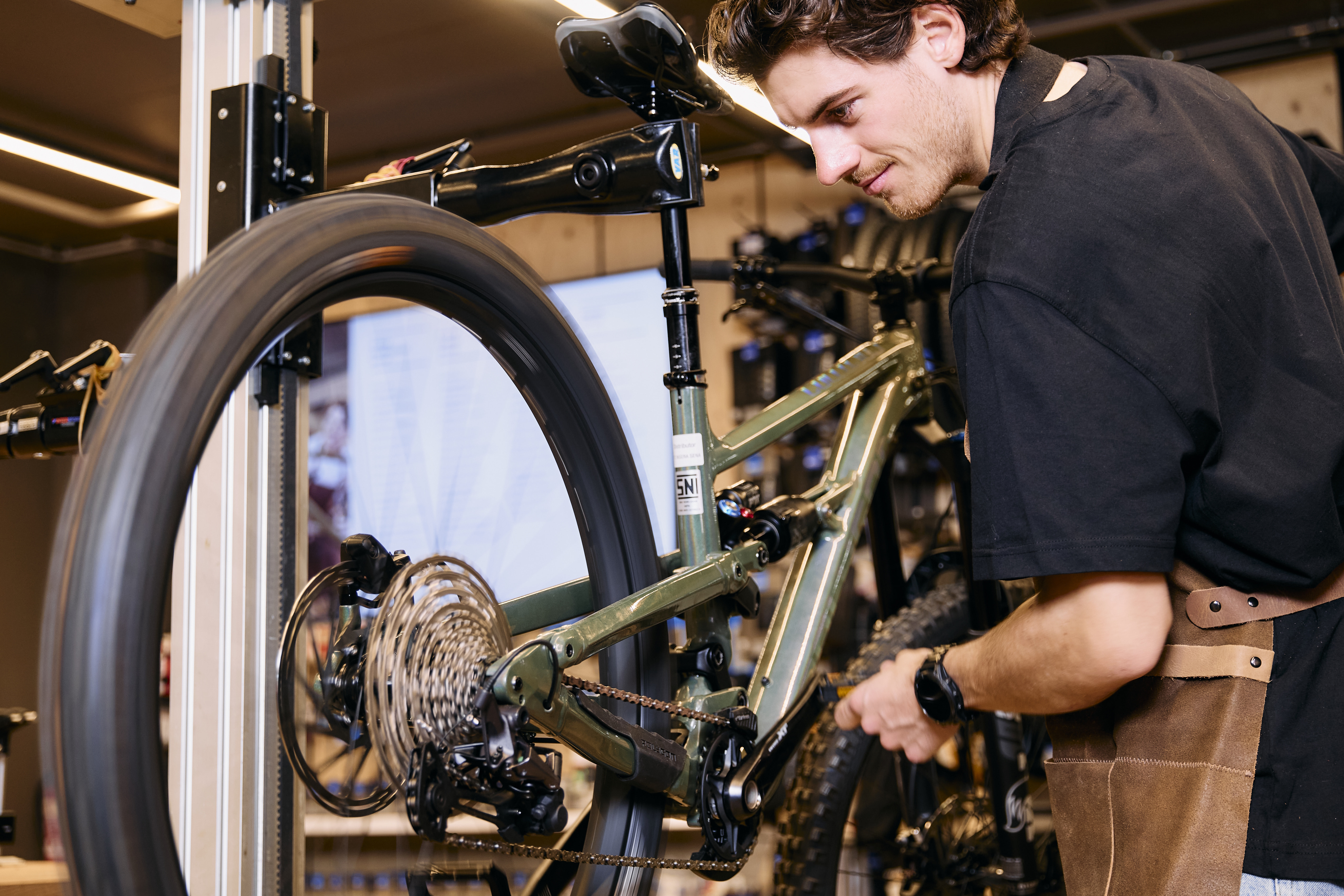 Ein konzentrierter Mechaniker in einer Werkstatt repariert ein grünes Mountainbike und justiert dessen Kette. Die Szene vermittelt Präzision und Hingabe in einem professionellen Umfeld.