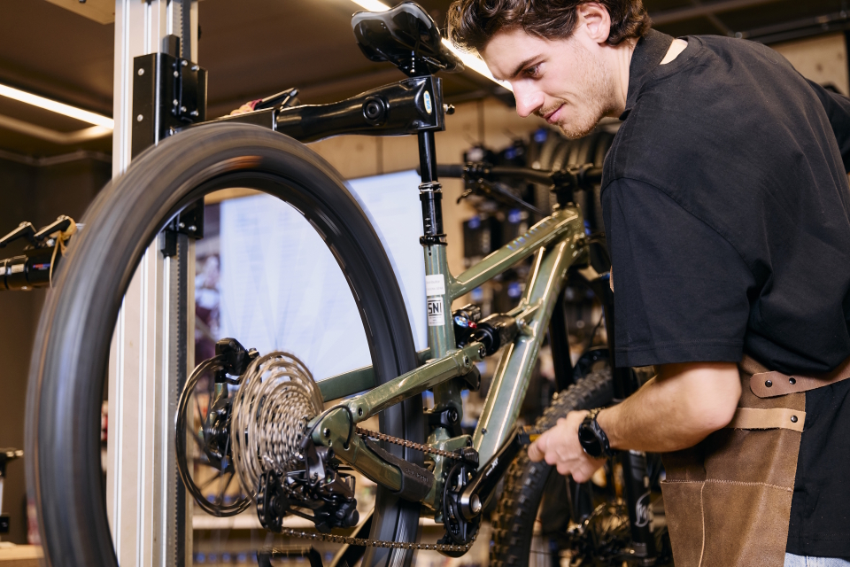 A focused mechanic in a workshop repairs a green mountain bike, adjusting its chain. The scene conveys precision and dedication in a professional setting.