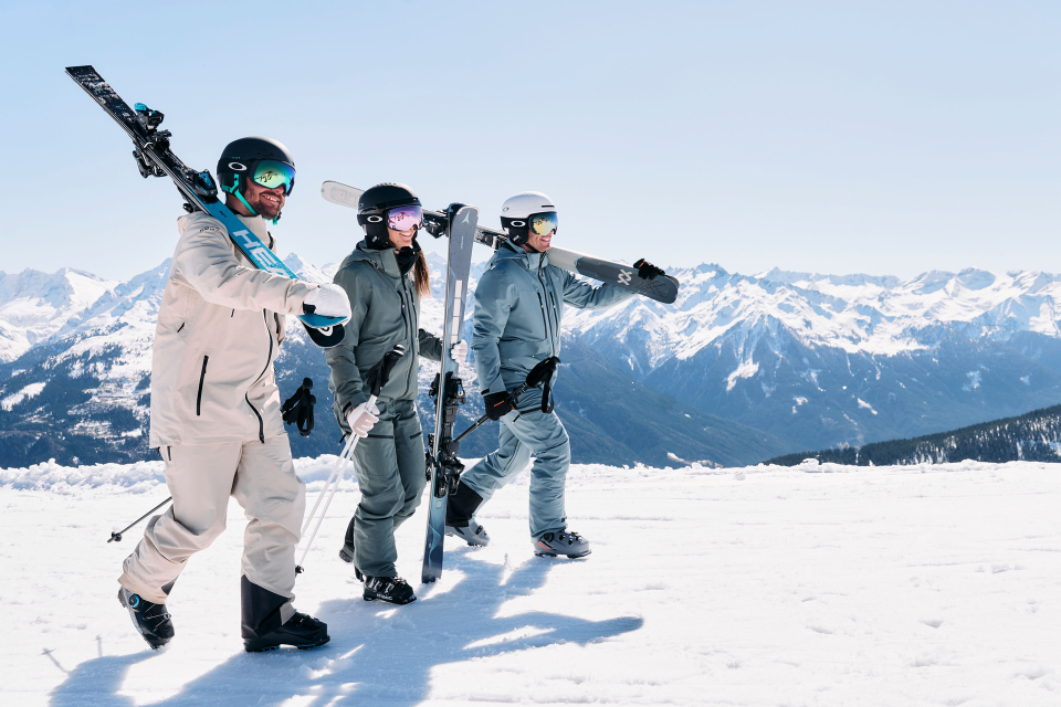 Three people in ski gear walk across a snowy mountain landscape, carrying skis. Bright, clear sky and majestic mountains in the background, conveying adventure.