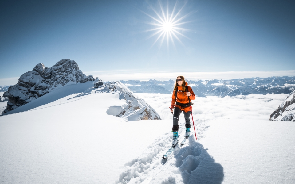 Vier Skifahrer fahren einen sonnenbeschienenen Schneehang hinunter und werfen dabei lange Schatten. Berge und ein klarer blauer Himmel bilden die Kulisse und vermitteln ein Gefühl von Abenteuer und Freude.