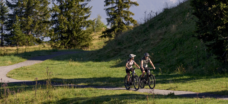 Zwei Radfahrer fahren mit Mountainbikes auf einem grasbewachsenen Weg mit malerischem Blick auf ferne Berge unter einem klaren blauen Himmel und vermitteln dabei Abenteuerlust und Freude.