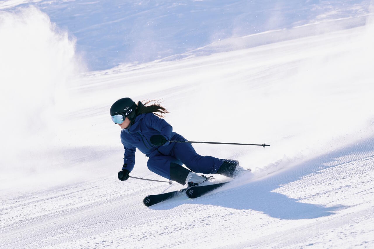 Ein Skifahrer in blauer Kleidung und mit Helm fährt schnell einen verschneiten Hang hinunter. Die Szene vermittelt Geschwindigkeit und Konzentration inmitten einer winterlichen Landschaft.