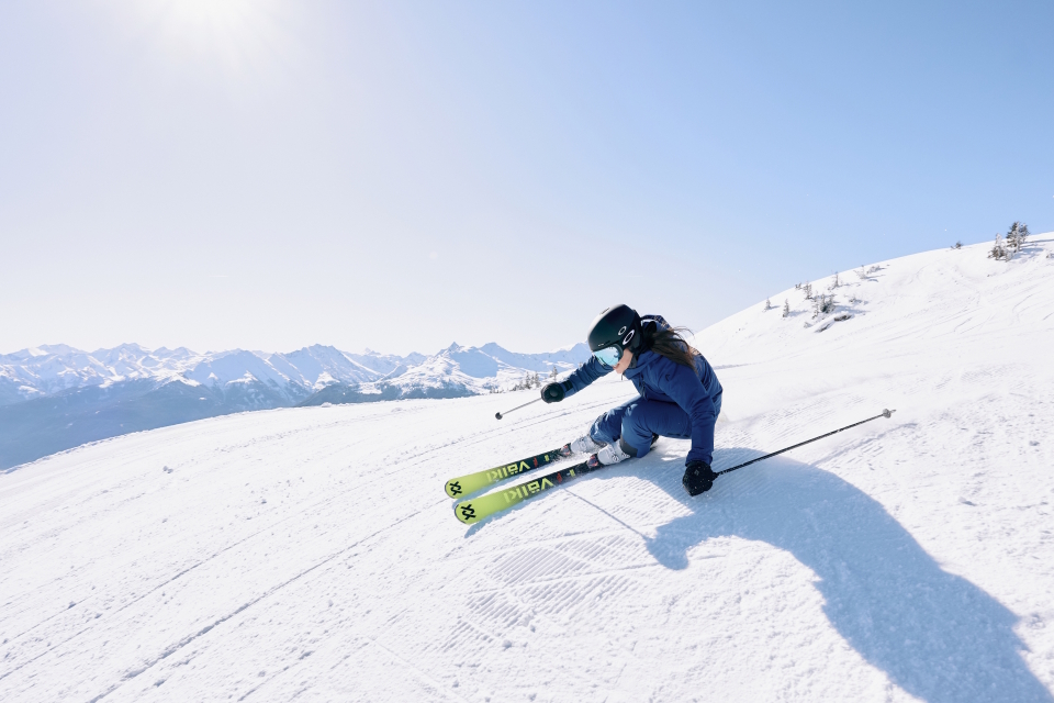 Skifahrerin in blauer Kleidung legt eine enge Carvingkurve auf sonniger Skipiste mit Alpenpanorama.