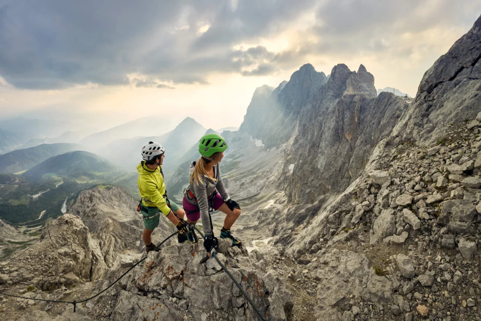 Two young hikers wearing helmets and outdoor gear stand on a rocky mountain ridge. The dramatic sky and distant peaks create a sense of adventure and awe.