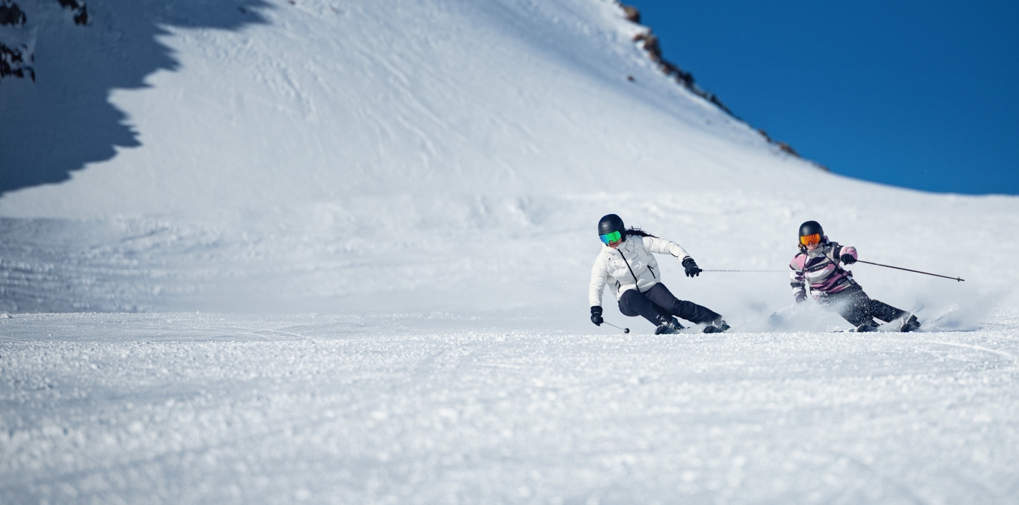Two skiers skiing downhill on a sunny day in a snowy mountain landscape.
