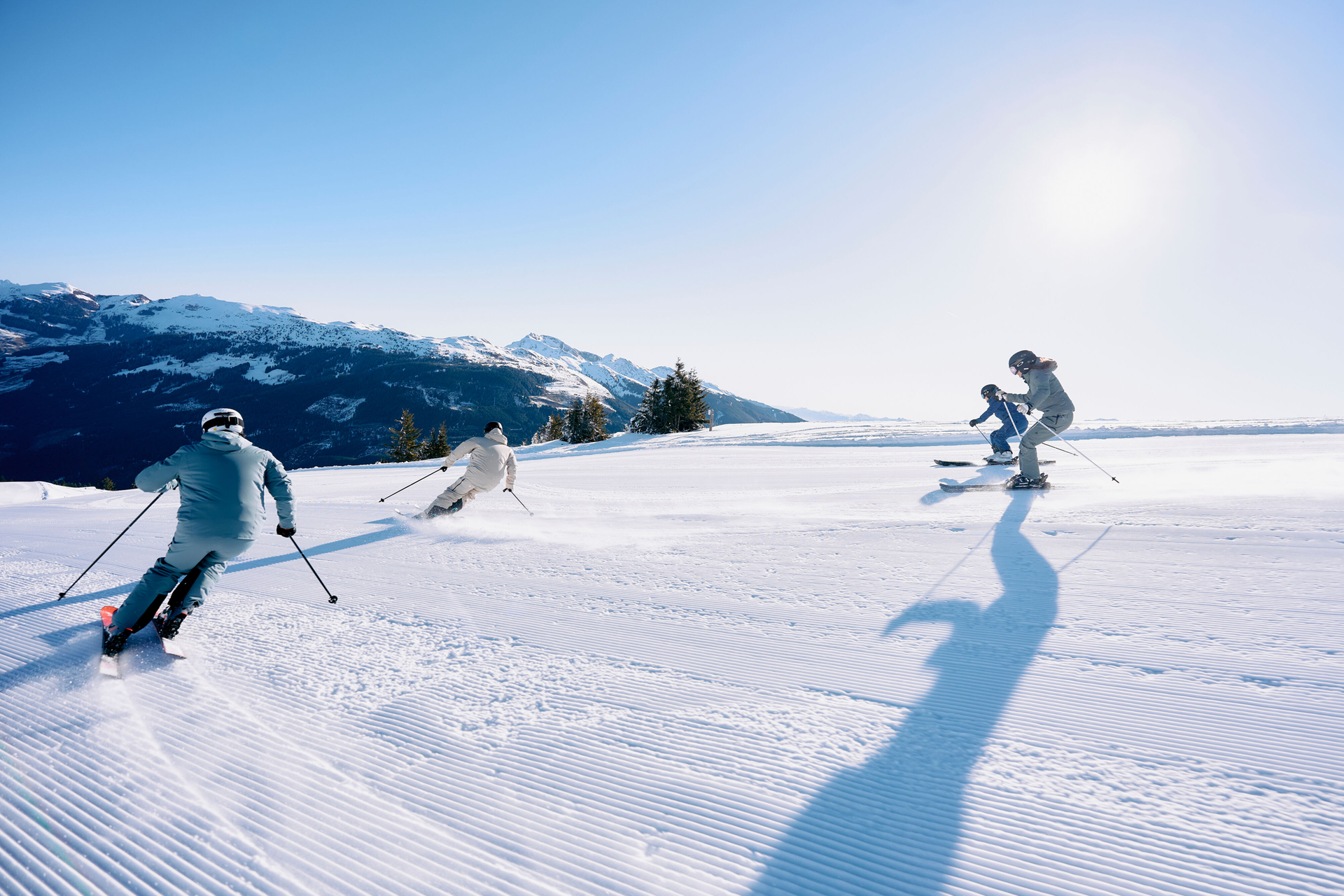 Vier Skifahrer fahren einen sonnenbeschienenen Schneehang hinunter und werfen dabei lange Schatten. Berge und ein klarer blauer Himmel bilden die Kulisse und vermitteln ein Gefühl von Abenteuer und Freude.