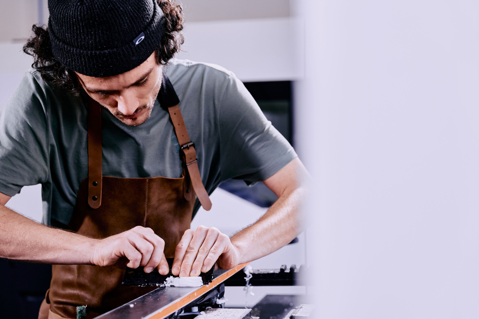 A man in a beanie and apron focuses intently on crafting or repairing an object, suggesting precision work. The setting is minimalist and modern.