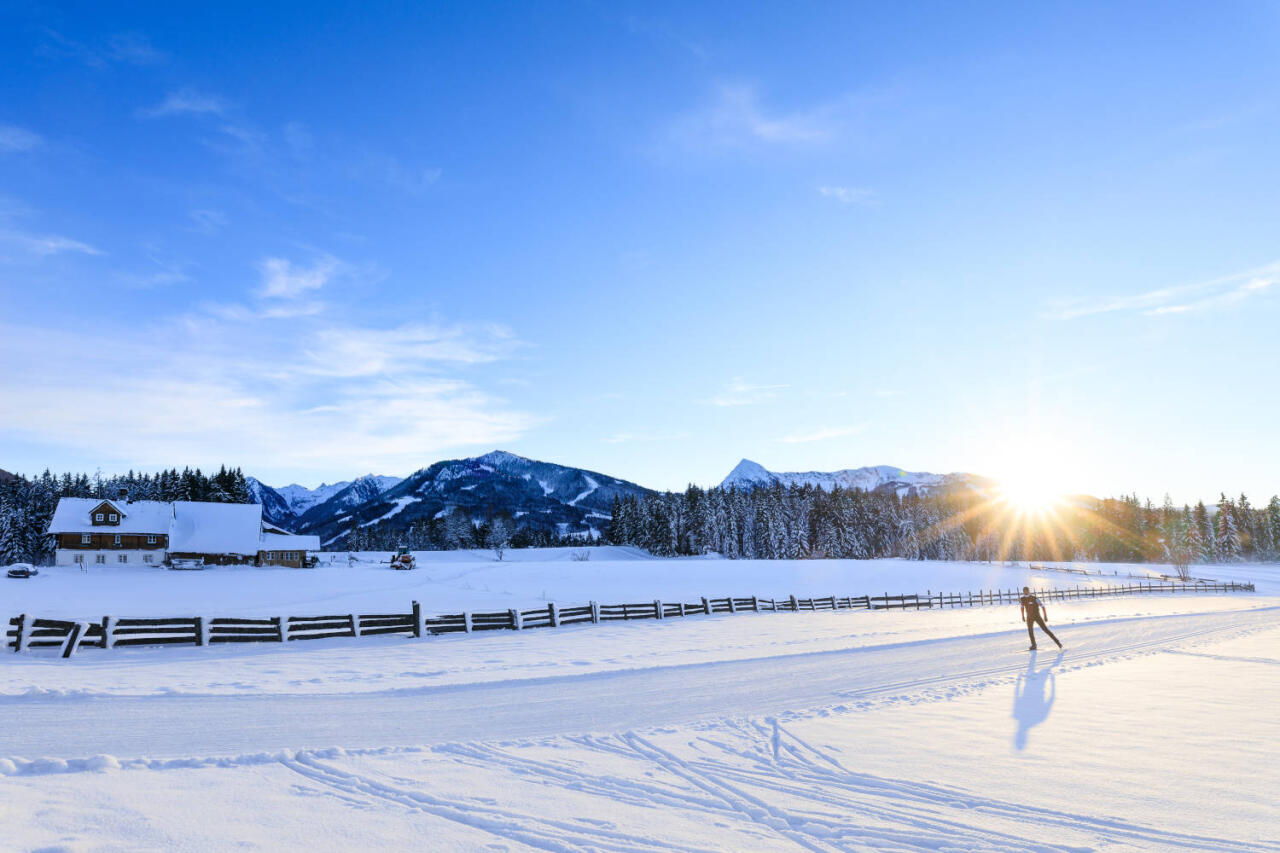 Langläufer gleitet bei Sonnenaufgang durch verschneite Winterlandschaft mit Bergen, Wald und Bauernhaus im Hintergrund.