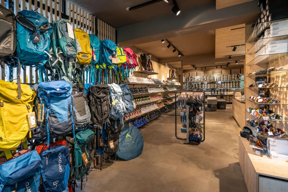 A well-lit outdoor gear store displays colorful backpacks on the left wall and various camping equipment on shelves. The ambiance is organized and inviting.