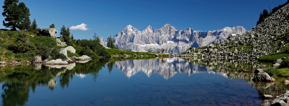 Clear mountain lake reflecting jagged peaks under a bright blue sky. Lush greenery and rocks line the shore, evoking a serene, natural beauty.