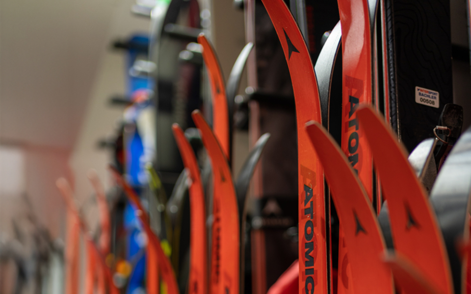 Cross-country skier glides through snow-covered winter landscape at sunrise with mountains, forest, and farmhouse in the background.